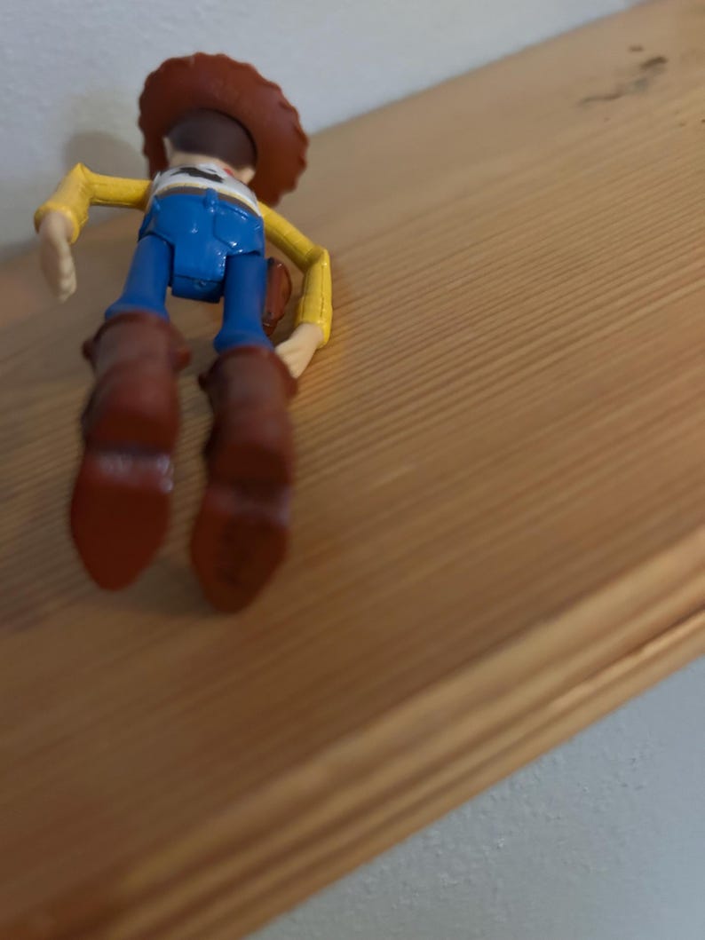 May include: A Woody action figure, viewed from the back, with a brown cowboy hat, yellow shirt, blue jeans, and brown boots. The figure is positioned on a wooden surface, showcasing the character's iconic outfit.