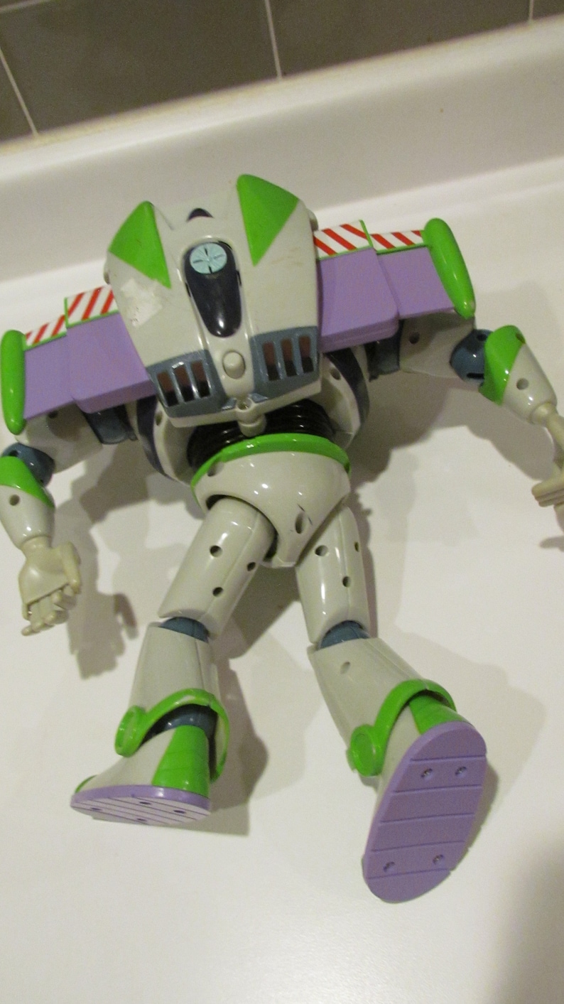 May include: A Buzz Lightyear action figure, featuring a white and green spacesuit with purple accents. The toy has articulated limbs and a jetpack with red and white stripes. The figure is posed on a white surface.