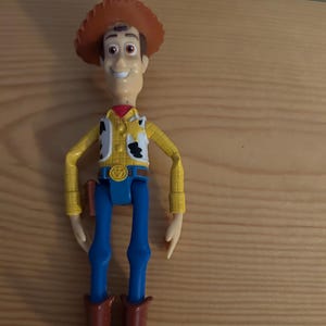 May include: A Woody action figure, featuring a brown cowboy hat, yellow shirt, white vest with black cow spots, blue pants, and brown boots. The figure has a smiling expression and a red neckerchief. A toy gun is in a holster.