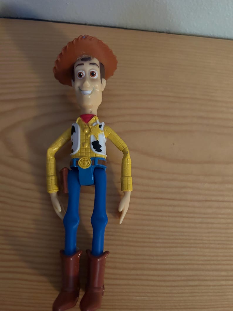 May include: A Woody action figure, featuring a brown cowboy hat, yellow shirt, white vest with black cow spots, blue pants, and brown boots. The figure has a smiling expression and a red neckerchief. A toy gun is in a holster.