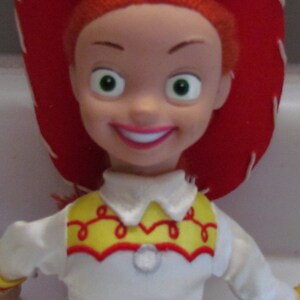 Pode incluir: Uma boneca Jessie da franquia Toy Story. A boneca tem cabelo ruivo, um chap&eacute;u de cowboy vermelho e uma camisa branca com detalhes amarelos e vermelhos. A boneca sorri com l&aacute;bios rosados e olhos verdes.