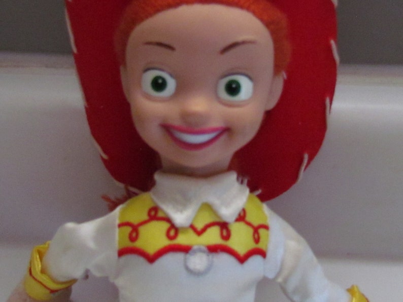 Pode incluir: Uma boneca Jessie da franquia Toy Story. A boneca tem cabelo ruivo, um chap&eacute;u de cowboy vermelho e uma camisa branca com detalhes amarelos e vermelhos. A boneca sorri com l&aacute;bios rosados e olhos verdes.