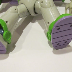 May include: Close-up of the feet of a Buzz Lightyear action figure. The figure's feet are light purple with green accents. The figure is wearing a white and green spacesuit.