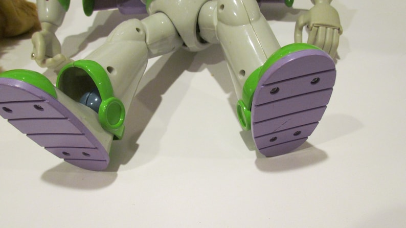 May include: Close-up of the feet of a Buzz Lightyear action figure. The figure's feet are light purple with green accents. The figure is wearing a white and green spacesuit.