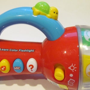 May include: A red and blue VTech Spin & Learn Color Flashlight toy with a handle and a small turtle on top. The toy has buttons with musical notes, ABCs, and a question mark. The flashlight has a clear rotating section with animal images.