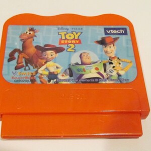 May include: Orange VTech SmartBook cartridge featuring characters from the Disney Pixar movie Toy Story 2. The cartridge has a picture of Woody, Buzz Lightyear, Jessie, and Bullseye. The text on the cartridge reads "Disney Pixar Toy Story 2" and "VTech".