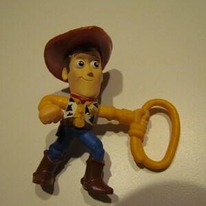 May include: A plastic toy figurine of Woody from Toy Story. He is wearing his signature brown cowboy hat, blue jeans, and a yellow vest with a red bandana. He is holding a yellow lasso in his right hand.
