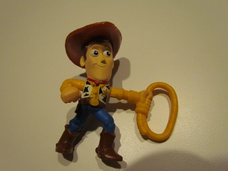 May include: A plastic toy figurine of Woody from Toy Story. He is wearing his signature brown cowboy hat, blue jeans, and a yellow vest with a red bandana. He is holding a yellow lasso in his right hand.
