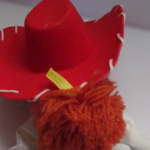 Pode incluir: Um close-up de um chap&eacute;u de cowboy vermelho com costuras brancas ao redor da aba. O chap&eacute;u est&aacute; sobre uma boneca com cabelo de fio laranja e uma fita amarela. A boneca est&aacute; vestindo uma camisa branca. Brinquedo.