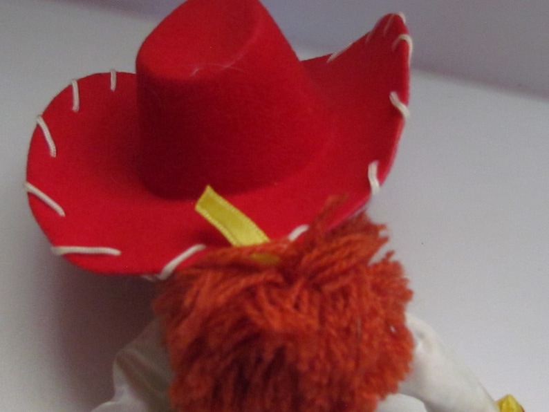 Pode incluir: Um close-up de um chap&eacute;u de cowboy vermelho com costuras brancas ao redor da aba. O chap&eacute;u est&aacute; sobre uma boneca com cabelo de fio laranja e uma fita amarela. A boneca est&aacute; vestindo uma camisa branca. Brinquedo.
