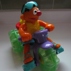 May include: Orange Sesame Street character riding a purple and green toy tricycle with a blue helmet and yellow life vest.