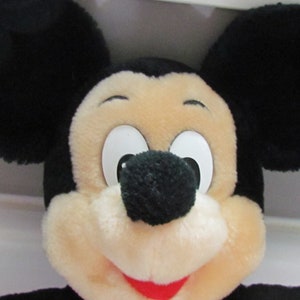 May include: Close-up of a plush Mickey Mouse toy. The toy features large black ears, a cream-colored face, and a black nose. The eyes are white with black pupils, and the mouth is red. The toy is a classic Disney character.