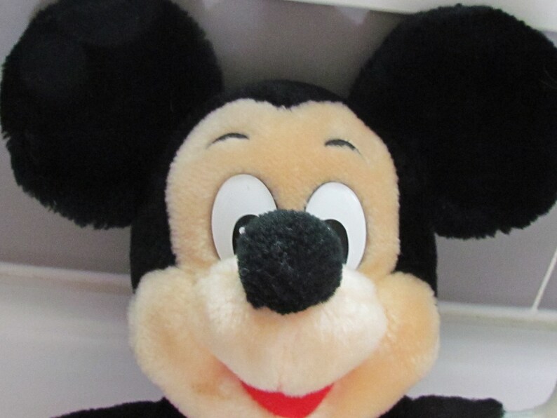 May include: Close-up of a plush Mickey Mouse toy. The toy features large black ears, a cream-colored face, and a black nose. The eyes are white with black pupils, and the mouth is red. The toy is a classic Disney character.