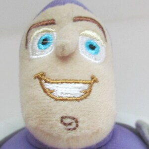 May include: Close-up of a plush toy's face. The toy has a purple hat, brown eyebrows, white eyes with blue pupils, a white smile with a gold outline, and a brown nose.