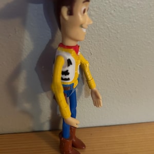 May include: A Woody action figure, featuring a brown cowboy hat, yellow shirt with a white vest, blue pants, and brown boots. The figure has a smiling expression and is posed in a standing position.