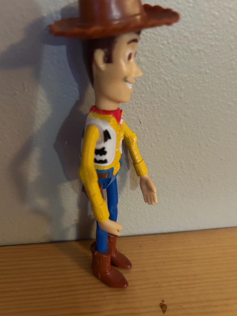 May include: A Woody action figure, featuring a brown cowboy hat, yellow shirt with a white vest, blue pants, and brown boots. The figure has a smiling expression and is posed in a standing position.