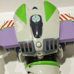 May include: A close-up of the back of a Buzz Lightyear action figure. The figure is primarily white, with green and purple accents. The wings are purple with red and white stripes. The figure has a button and a small light.