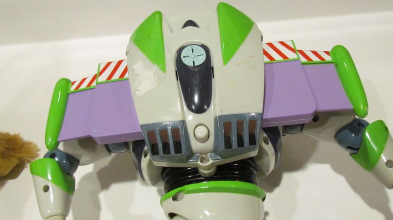 May include: A close-up of the back of a Buzz Lightyear action figure. The figure is primarily white, with green and purple accents. The wings are purple with red and white stripes. The figure has a button and a small light.