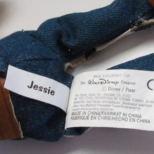 Pode incluir: Close-up do tecido semelhante a ganga e dos detalhes castanhos de um brinquedo de pel&uacute;cia. Uma etiqueta branca diz "Jessie" em texto preto. Outra etiqueta indica que o brinquedo &eacute; feito exclusivamente para The Walt Disney Company.