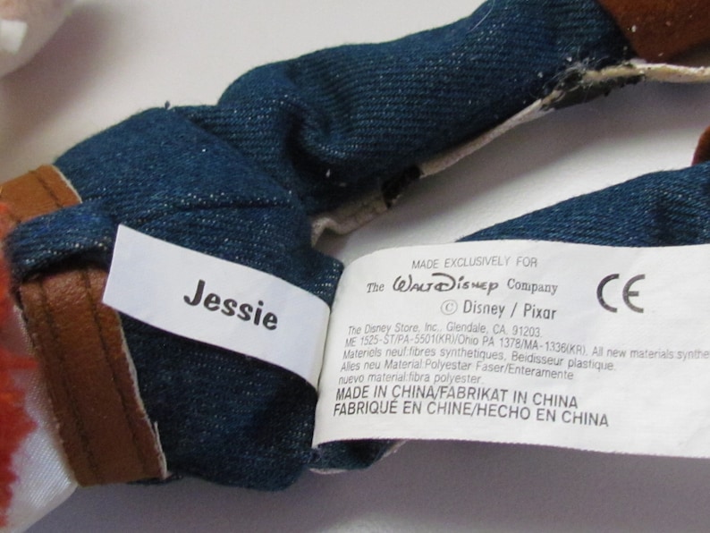 Pode incluir: Close-up do tecido semelhante a ganga e dos detalhes castanhos de um brinquedo de pel&uacute;cia. Uma etiqueta branca diz "Jessie" em texto preto. Outra etiqueta indica que o brinquedo &eacute; feito exclusivamente para The Walt Disney Company.