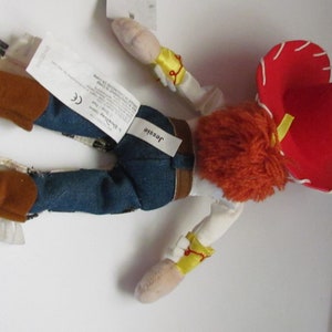 Pode incluir: Uma boneca de pel&uacute;cia Jessie da franquia Toy Story. A boneca est&aacute; usando um chap&eacute;u vermelho, jeans azuis e botas marrons. A boneca tem cabelo de fio laranja e um colete amarelo. O nome "Jessie" est&aacute; impresso em uma etiqueta.
