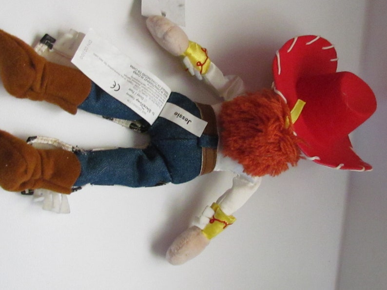 Pode incluir: Uma boneca de pel&uacute;cia Jessie da franquia Toy Story. A boneca est&aacute; usando um chap&eacute;u vermelho, jeans azuis e botas marrons. A boneca tem cabelo de fio laranja e um colete amarelo. O nome "Jessie" est&aacute; impresso em uma etiqueta.