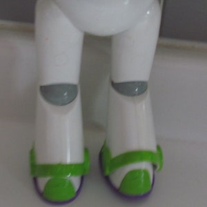 Puede incluir: Primer plano de la parte inferior del cuerpo de una figura de acci&oacute;n de Buzz Lightyear. Las piernas blancas de la figura tienen rodilleras grises y detalles verdes y morados en los pies. La figura est&aacute; sobre una superficie blanca.