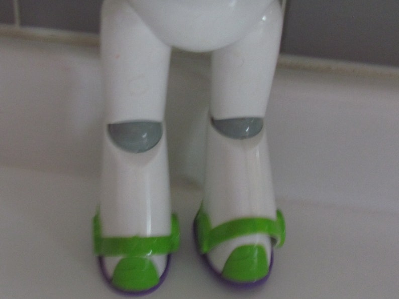 Puede incluir: Primer plano de la parte inferior del cuerpo de una figura de acci&oacute;n de Buzz Lightyear. Las piernas blancas de la figura tienen rodilleras grises y detalles verdes y morados en los pies. La figura est&aacute; sobre una superficie blanca.