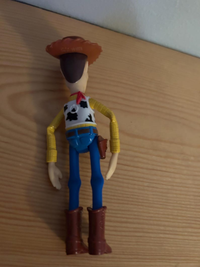May include: A Woody action figure, featuring a brown cowboy hat, a white vest with a black and white cow pattern, a red neckerchief, yellow sleeves, blue pants, and brown boots. A toy gun is in a holster on the right side.