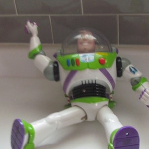 Puede incluir: Figura de acci&oacute;n de Buzz Lightyear en posici&oacute;n sentada. El juguete presenta un traje espacial blanco y verde con detalles morados. La figura tiene un casco transparente y es un art&iacute;culo de colecci&oacute;n de la franquicia Toy Story.