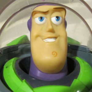 May include: A close-up of a Buzz Lightyear action figure. The toy has a green and purple space suit, a clear plastic helmet, and a smiling face with blue eyes. The figure is a popular character from the Toy Story franchise.