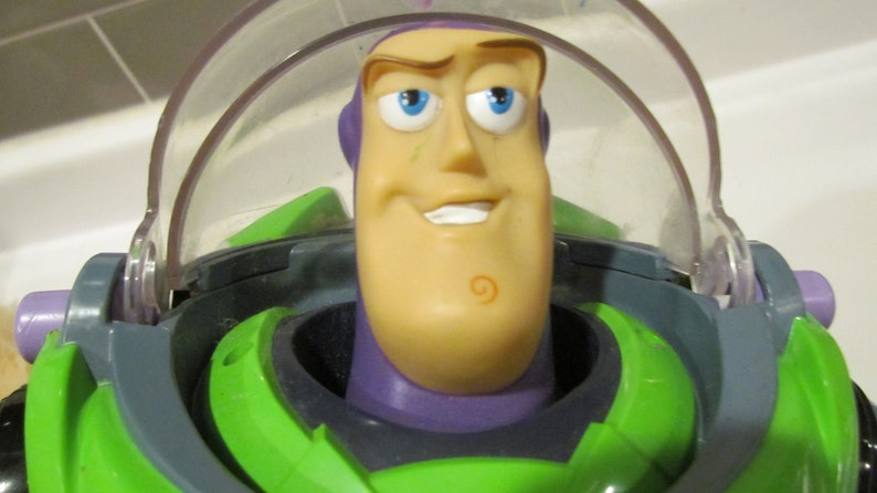 May include: A close-up of a Buzz Lightyear action figure. The toy has a green and purple space suit, a clear plastic helmet, and a smiling face with blue eyes. The figure is a popular character from the Toy Story franchise.