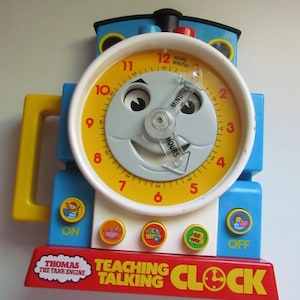 May include: A colorful Thomas the Tank Engine teaching clock. The clock face is yellow with black numbers and a smiling face. The clock has blue and red accents, with buttons labeled "ON" and "OFF". The text "TEACHING TALKING CLOCK" is visible.