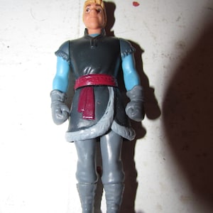 K&ouml;nnte beinhalten: Eine Plastikfigur einer Figur aus dem Animationsfilm "Die Eisk&ouml;nigin". Die Figur tr&auml;gt eine dunkelgraue Tunika mit hellblauen &Auml;rmeln, grauen Handschuhen und grauer Hose. Ein roter Sch&auml;rpe und ein pelzbesetzter Saum sorgen f&uuml;r Details.