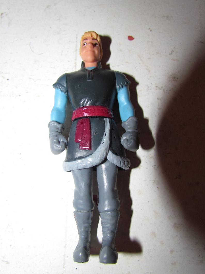 K&ouml;nnte beinhalten: Eine Plastikfigur einer Figur aus dem Animationsfilm "Die Eisk&ouml;nigin". Die Figur tr&auml;gt eine dunkelgraue Tunika mit hellblauen &Auml;rmeln, grauen Handschuhen und grauer Hose. Ein roter Sch&auml;rpe und ein pelzbesetzter Saum sorgen f&uuml;r Details.