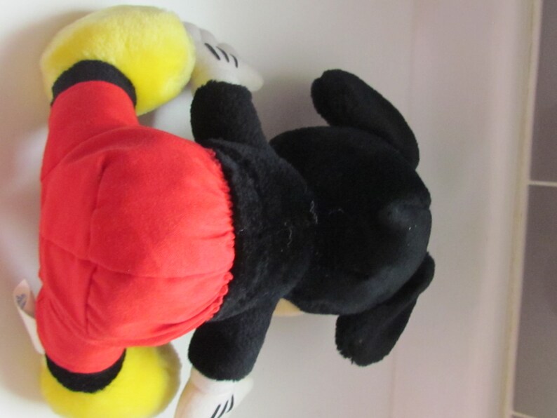 May include: A plush Mickey Mouse toy, featuring a red shorts, black body, yellow shoes, and white gloves. The toy is lying on its side, showcasing its iconic design and colors. A classic Disney character.