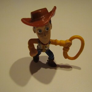May include: A plastic toy figurine of Woody from Toy Story. He is wearing a brown cowboy hat, a yellow shirt, and blue jeans. He is holding a yellow lasso in his right hand.