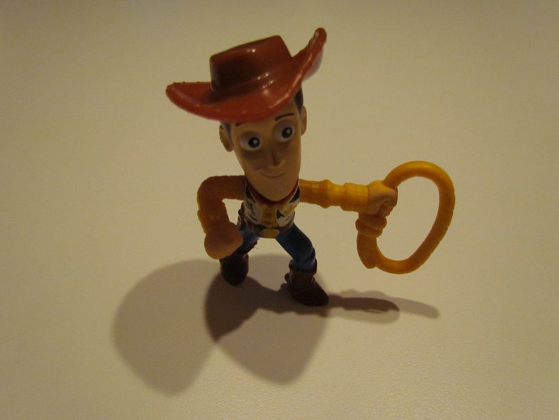 May include: A plastic toy figurine of Woody from Toy Story. He is wearing a brown cowboy hat, a yellow shirt, and blue jeans. He is holding a yellow lasso in his right hand.