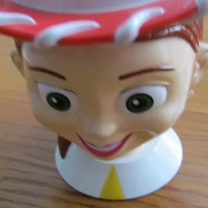 May include: A red and white cowboy hat with a smiling face and brown hair. The hat is made of plastic and has a yellow accent on the front.