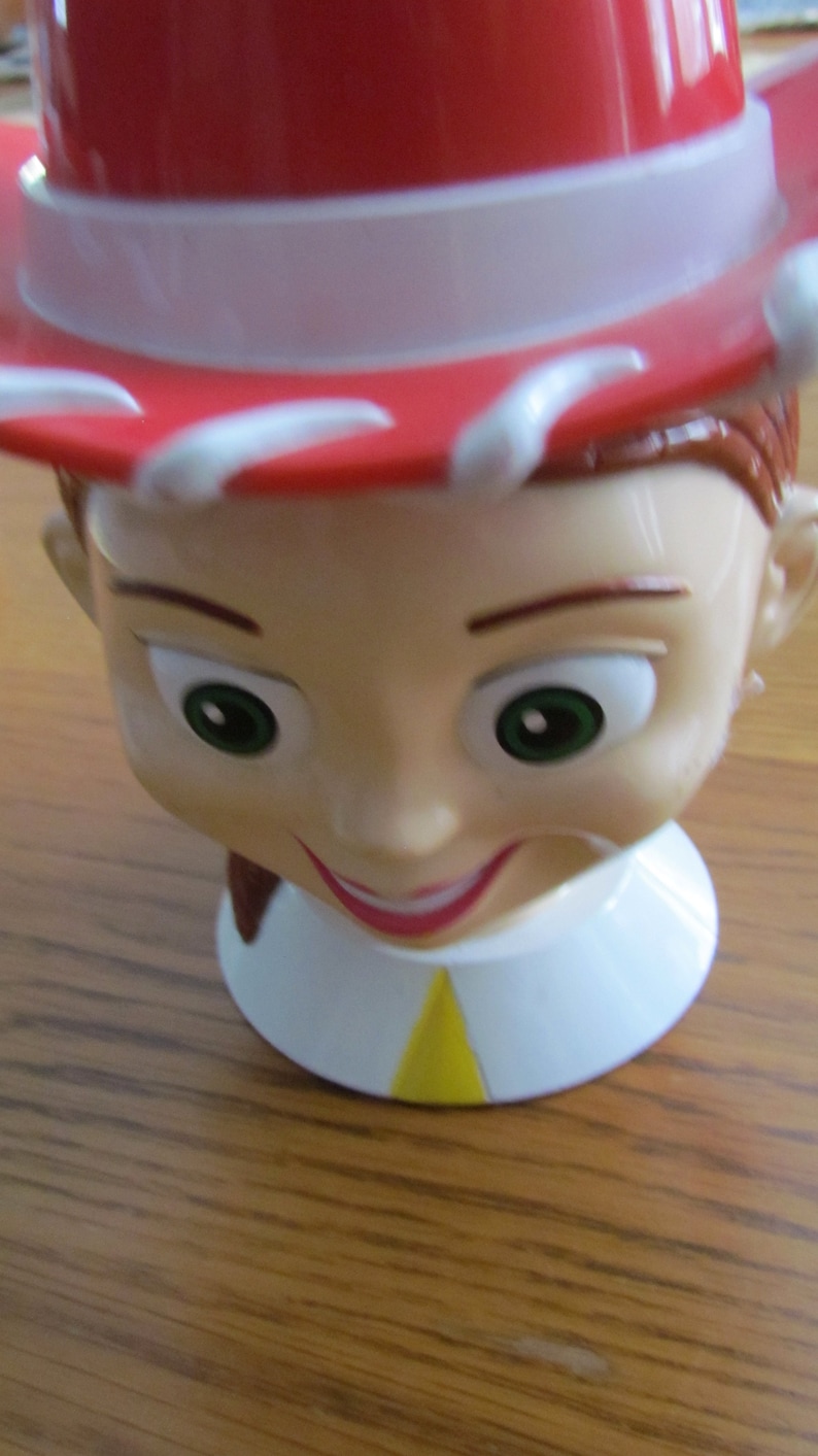 May include: A red and white cowboy hat with a smiling face and brown hair. The hat is made of plastic and has a yellow accent on the front.