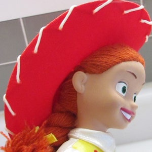 Pode incluir: Um close-up de uma boneca Jessie da franquia Toy Story. A boneca tem cabelo ruivo em uma tran&ccedil;a, um chap&eacute;u vermelho com costuras brancas e uma camisa branca com detalhes amarelos e vermelhos. A boneca est&aacute; sorrindo.