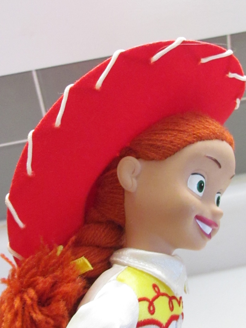 Pode incluir: Um close-up de uma boneca Jessie da franquia Toy Story. A boneca tem cabelo ruivo em uma tran&ccedil;a, um chap&eacute;u vermelho com costuras brancas e uma camisa branca com detalhes amarelos e vermelhos. A boneca est&aacute; sorrindo.