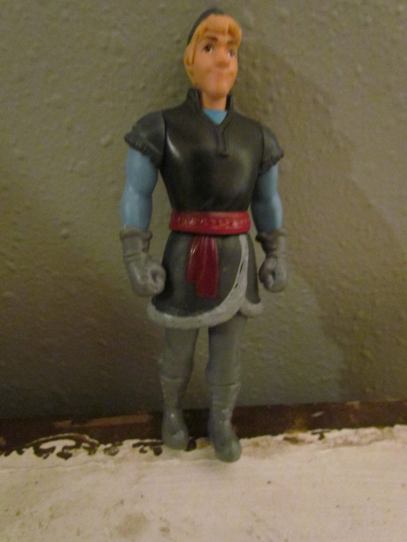 K&ouml;nnte beinhalten: Eine Plastikfigur einer Figur aus dem Animationsfilm "Die Eisk&ouml;nigin". Die Figur tr&auml;gt eine dunkelgraue Tunika mit blauen &Auml;rmeln, einen roten G&uuml;rtel und graue Hosen. Die Figur hat blondes Haar und steht aufrecht.