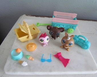 May include: A collection of colorful miniature toys, including animal figures, a picnic basket, a wagon, and food items. The toys are arranged on a white surface, with a light background.