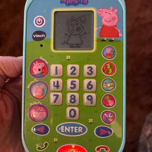 May include: A teal and green toy phone featuring Peppa Pig characters. The phone has a small screen displaying a pixelated image of Peppa Pig, a number pad, and character buttons. The phone has the VTech logo and the text "Peppa Pig".