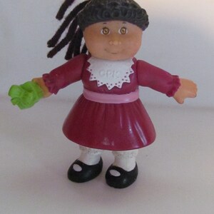 May include: A vintage Cabbage Patch Kids doll with dark hair in pigtails and a pink bow. The doll wears a burgundy dress, white bib with "CPK" text, black shoes, and a green glove. A classic collectible toy.
