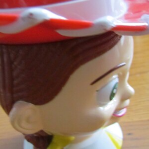 May include: A red and white cowboy hat with a brown hairpiece and a white face with green eyes and a pink mouth. The face is on a white base with a yellow and brown accent.