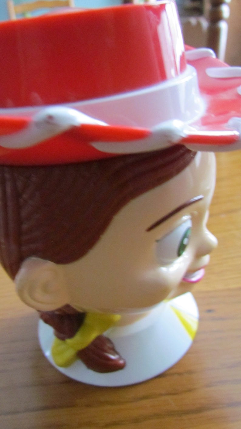 May include: A red and white cowboy hat with a brown hairpiece and a white face with green eyes and a pink mouth. The face is on a white base with a yellow and brown accent.