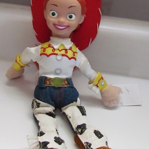 Pode incluir: Uma boneca Jessie da franquia Toy Story. A boneca tem um chap&eacute;u vermelho, cabelo laranja e uma camisa branca com detalhes amarelos e vermelhos. Ela usa cal&ccedil;as jeans azuis, um cinto marrom e chaps brancos com manchas pretas. Suas botas s&atilde;o marrons.