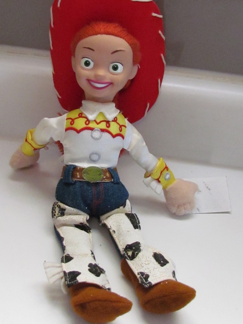 Pode incluir: Uma boneca Jessie da franquia Toy Story. A boneca tem um chap&eacute;u vermelho, cabelo laranja e uma camisa branca com detalhes amarelos e vermelhos. Ela usa cal&ccedil;as jeans azuis, um cinto marrom e chaps brancos com manchas pretas. Suas botas s&atilde;o marrons.
